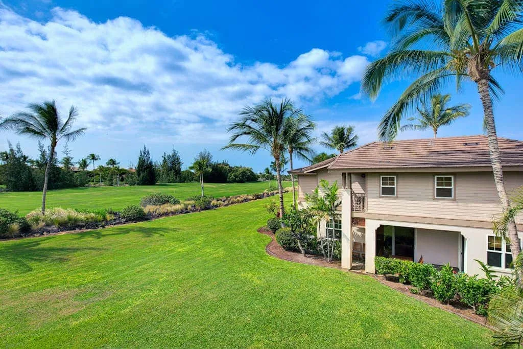 A two-story house stands beside a broad green lawn and palm trees.