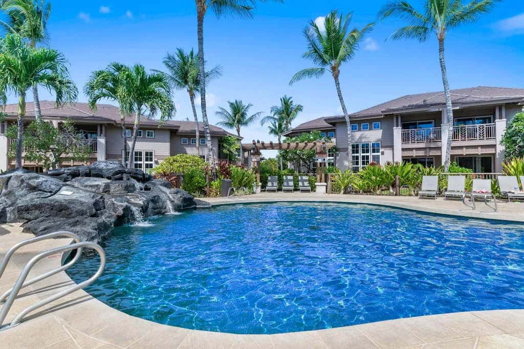 A swimming pool sits among palm trees and low-rise residential buildings.