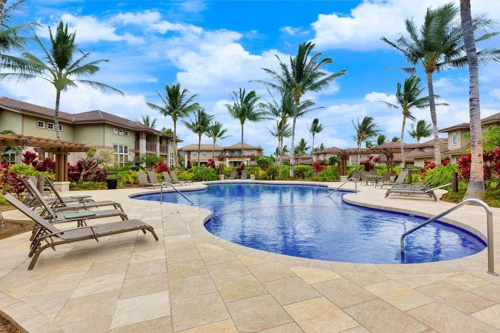A swimming pool is surrounded by lounge seating, palm trees, and residential buildings.