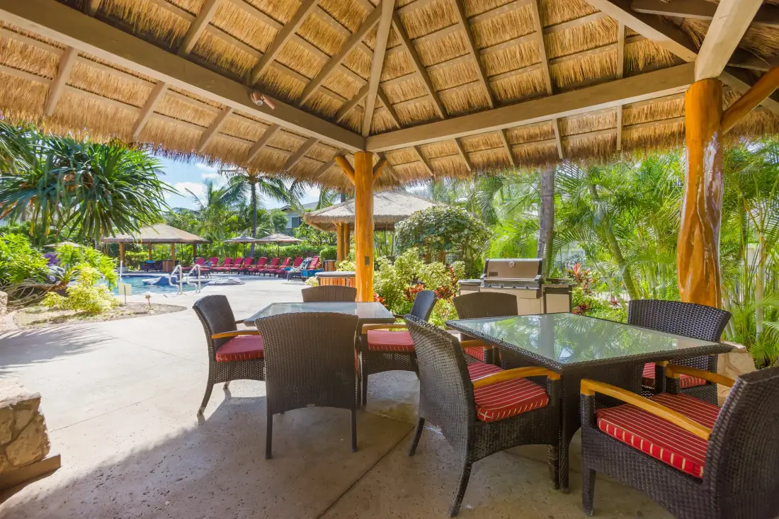 A covered outdoor dining area with wicker seating and a table overlooks a pool.