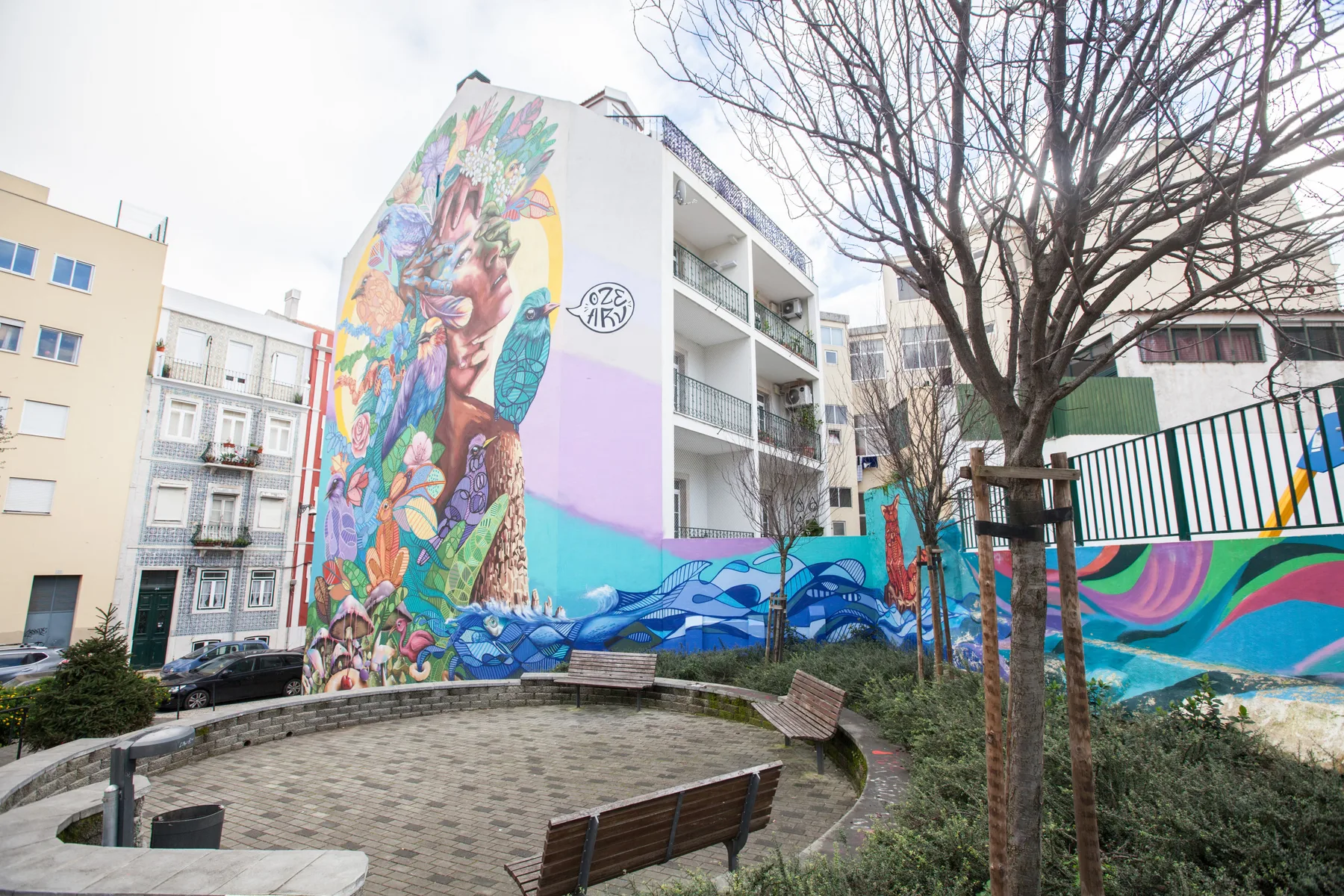 An urban courtyard features a large colorful mural and benches.