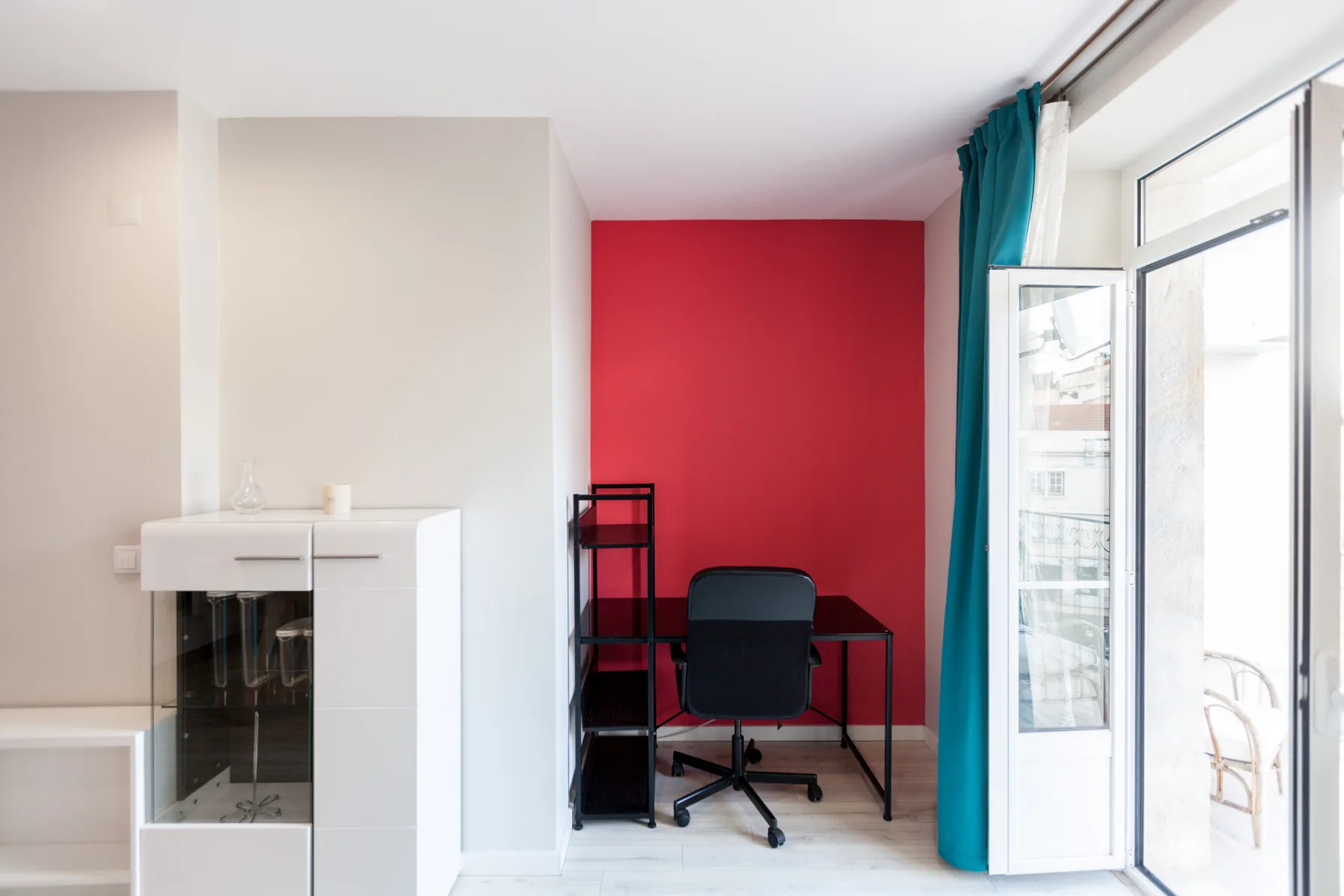 A small workspace with a desk and chair stands against a red wall beside a glass door.