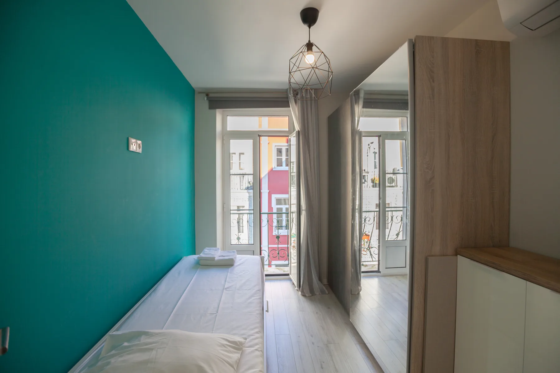 A narrow bedroom with a single bed beside a teal wall and tall windows.