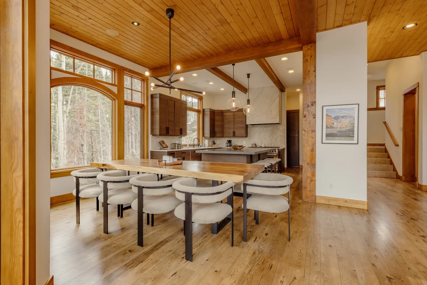 A dining area with a long table sits beside a kitchen and large windows.