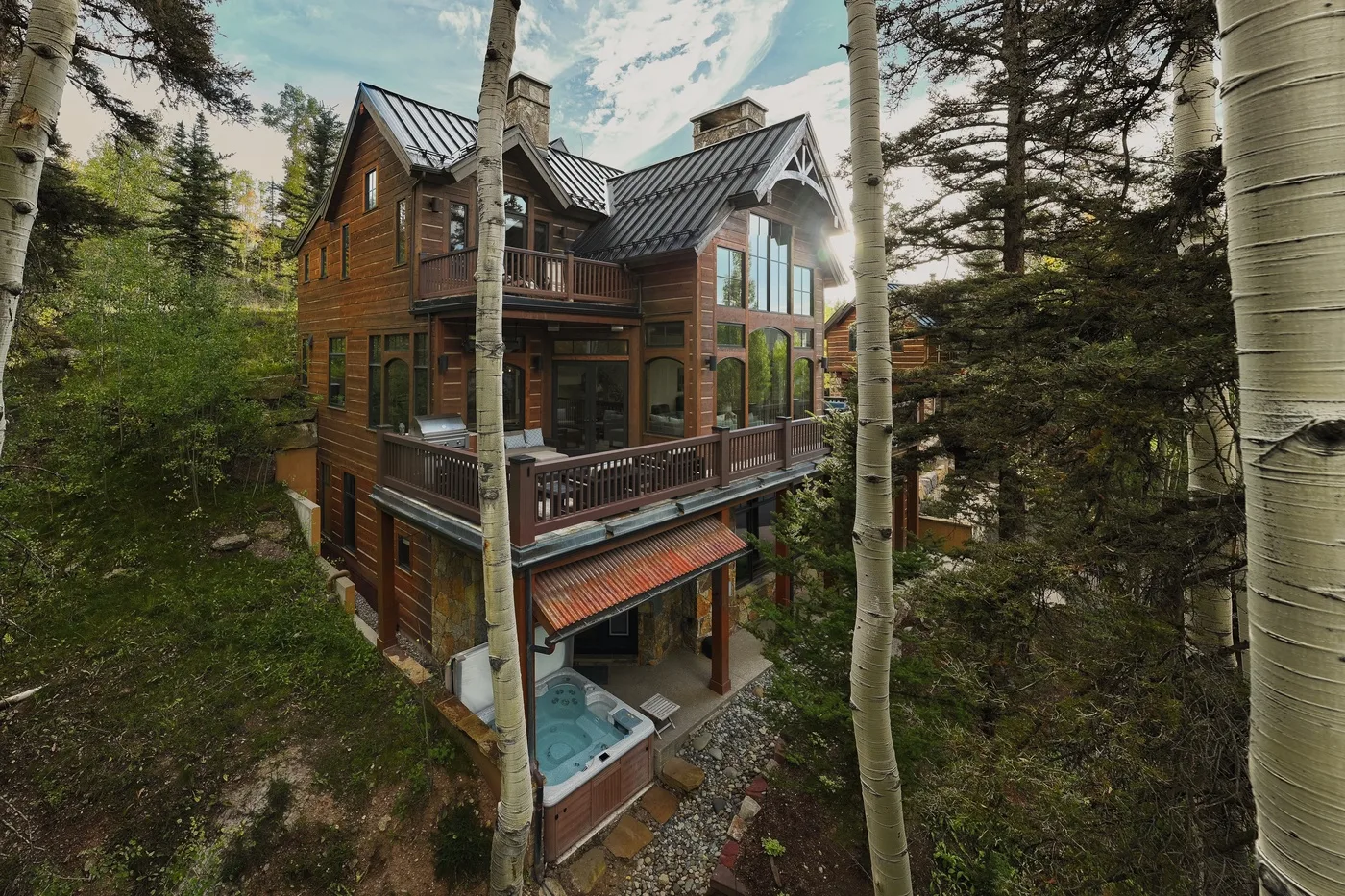 A multi-level wooden house sits among trees, with a hot tub beside the lower patio.