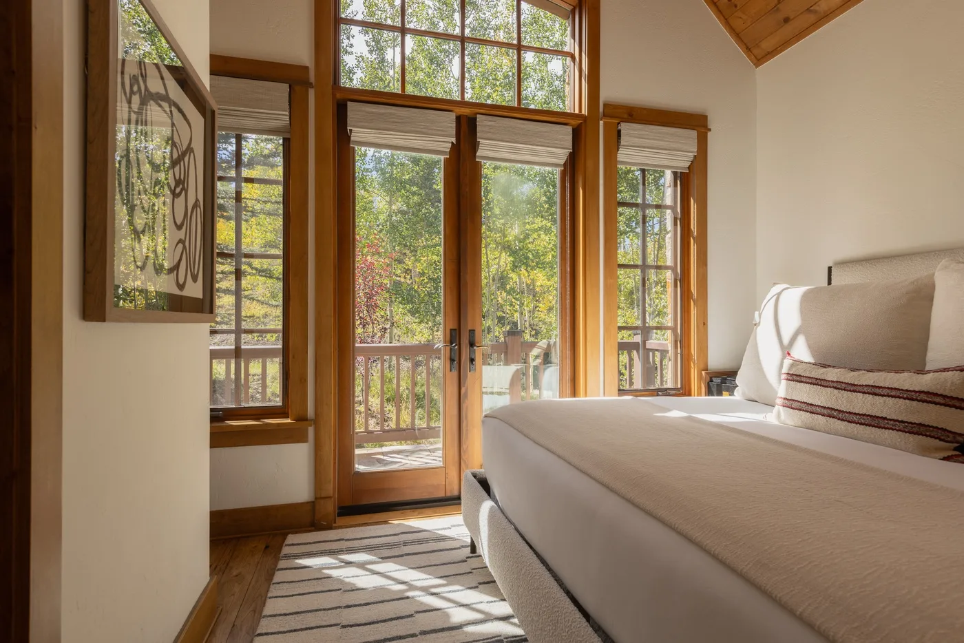 A bright bedroom with a bed beside tall windows overlooking leafy trees.