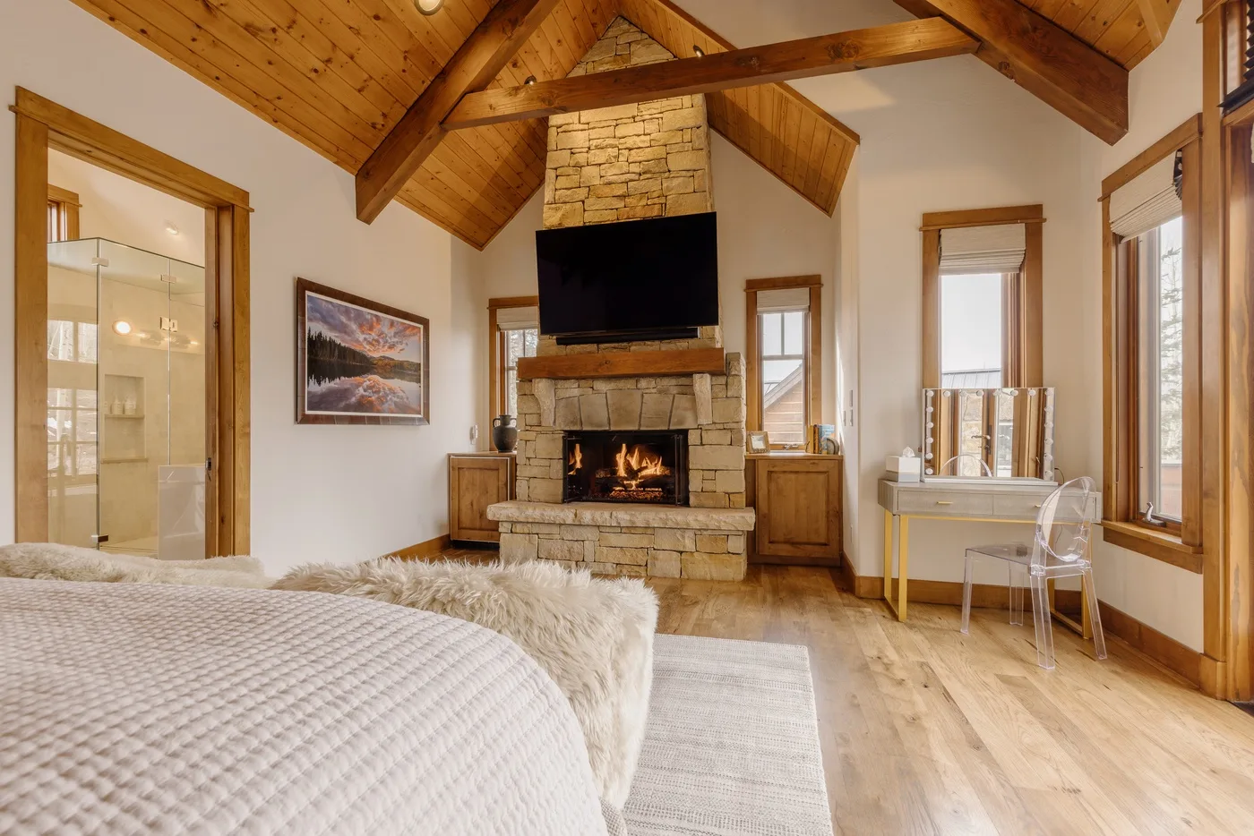 A bedroom with a stone fireplace, television, and sloped wooden ceiling.