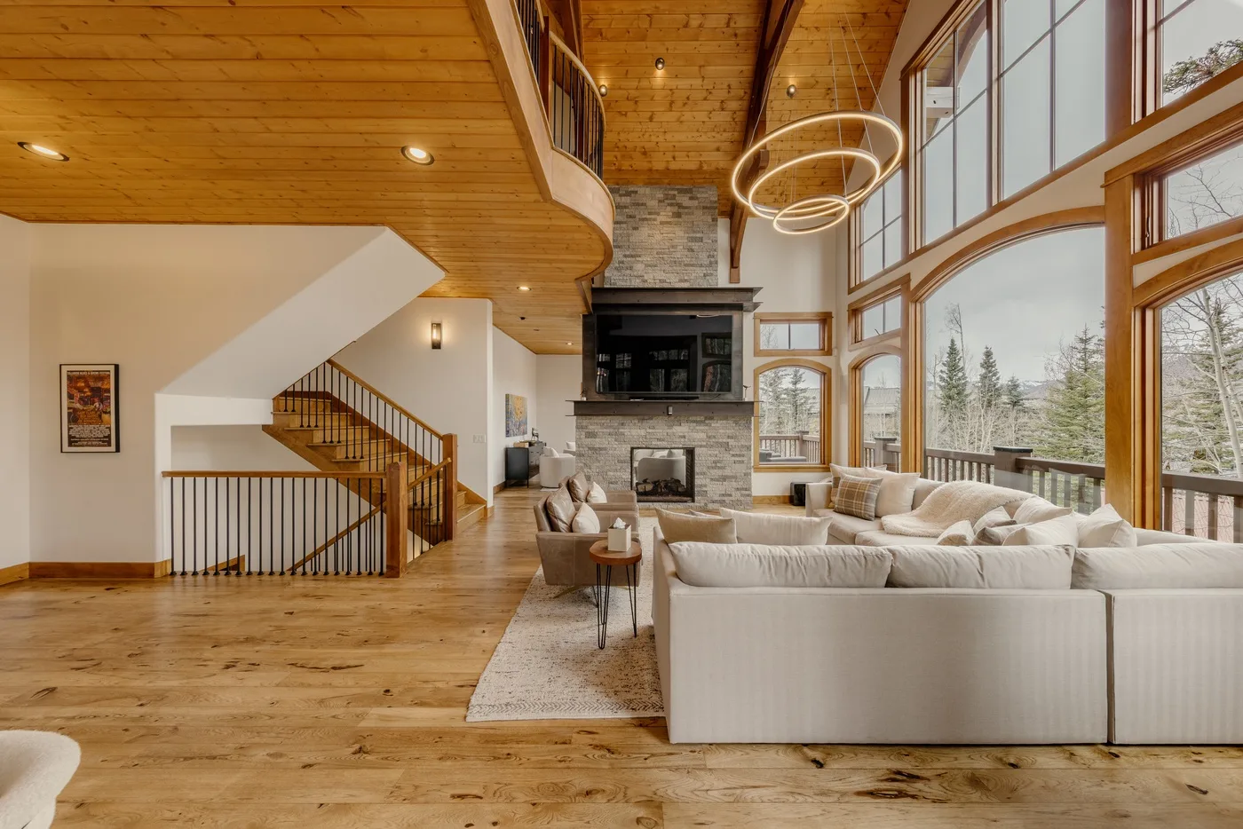 A spacious living room has light seating beside a stone fireplace and tall windows.