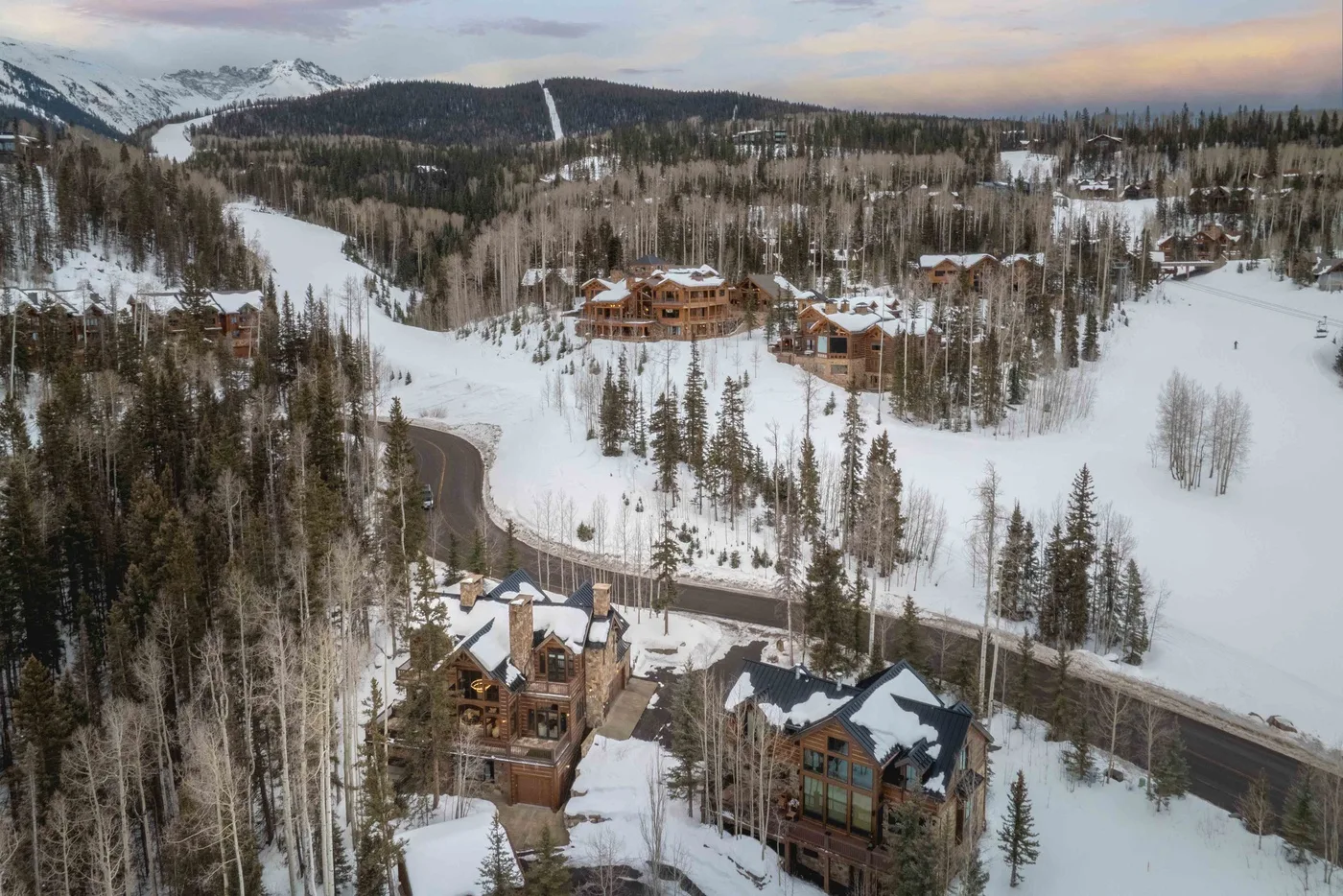 Snow-covered mountain landscape with wooded slopes, scattered buildings, and a winding road.