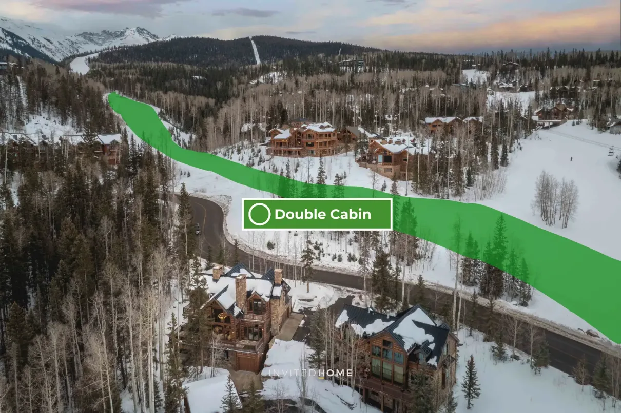 Snow-covered cabins sit among trees beside a winding road, with mountains in the distance.
