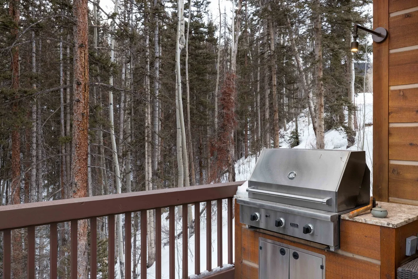 A wooden deck with a metal grill overlooks a snow-dusted forest.