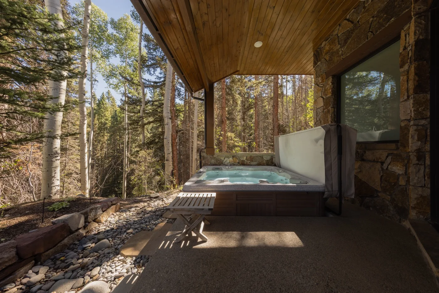 A covered outdoor hot tub sits beside a stone wall, overlooking a wooded setting.