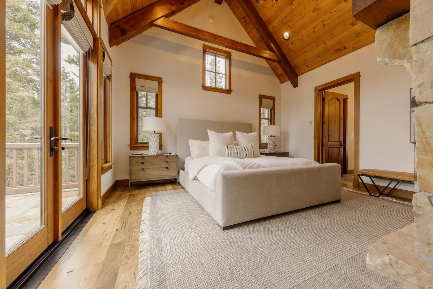 A bedroom with a bed beneath a vaulted wooden ceiling and windows overlooking the trees.