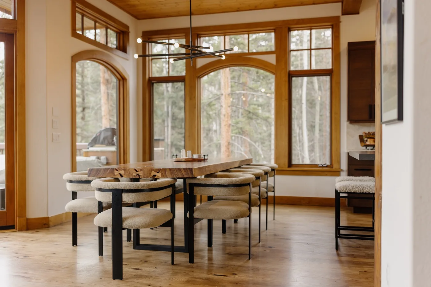 A dining area with a long table and large windows overlooking trees.