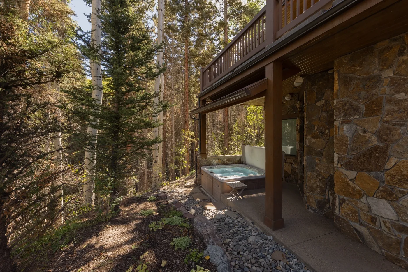 A wooded outdoor hot tub sits beside a stone-clad building.