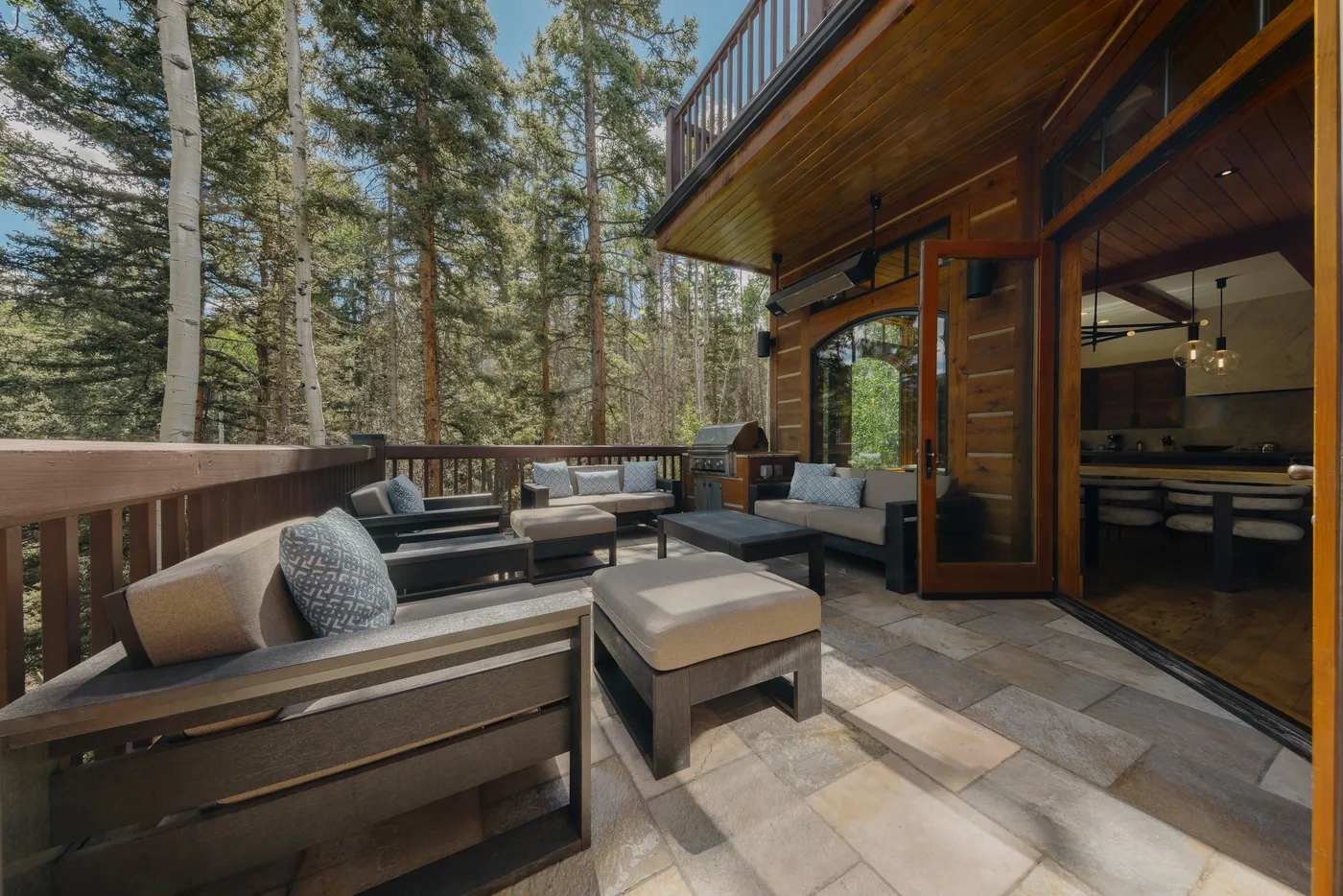 A furnished outdoor patio beside a wooden house, surrounded by tall trees.