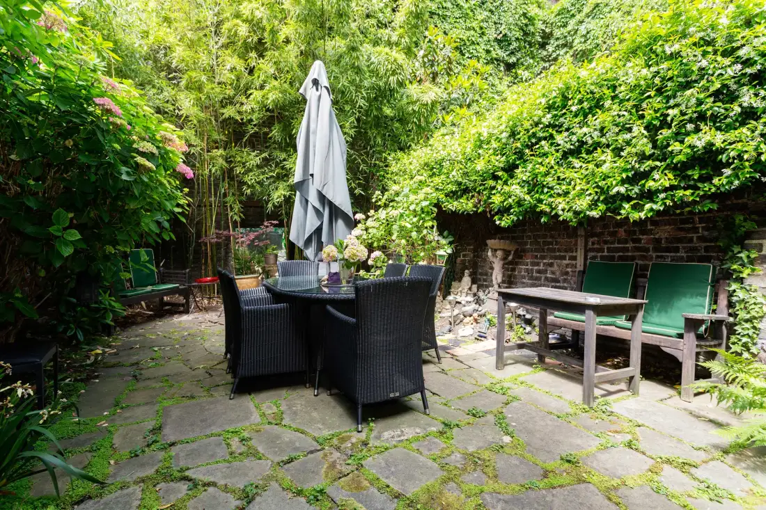 A leafy outdoor patio with a dining table, seating, and a closed umbrella.