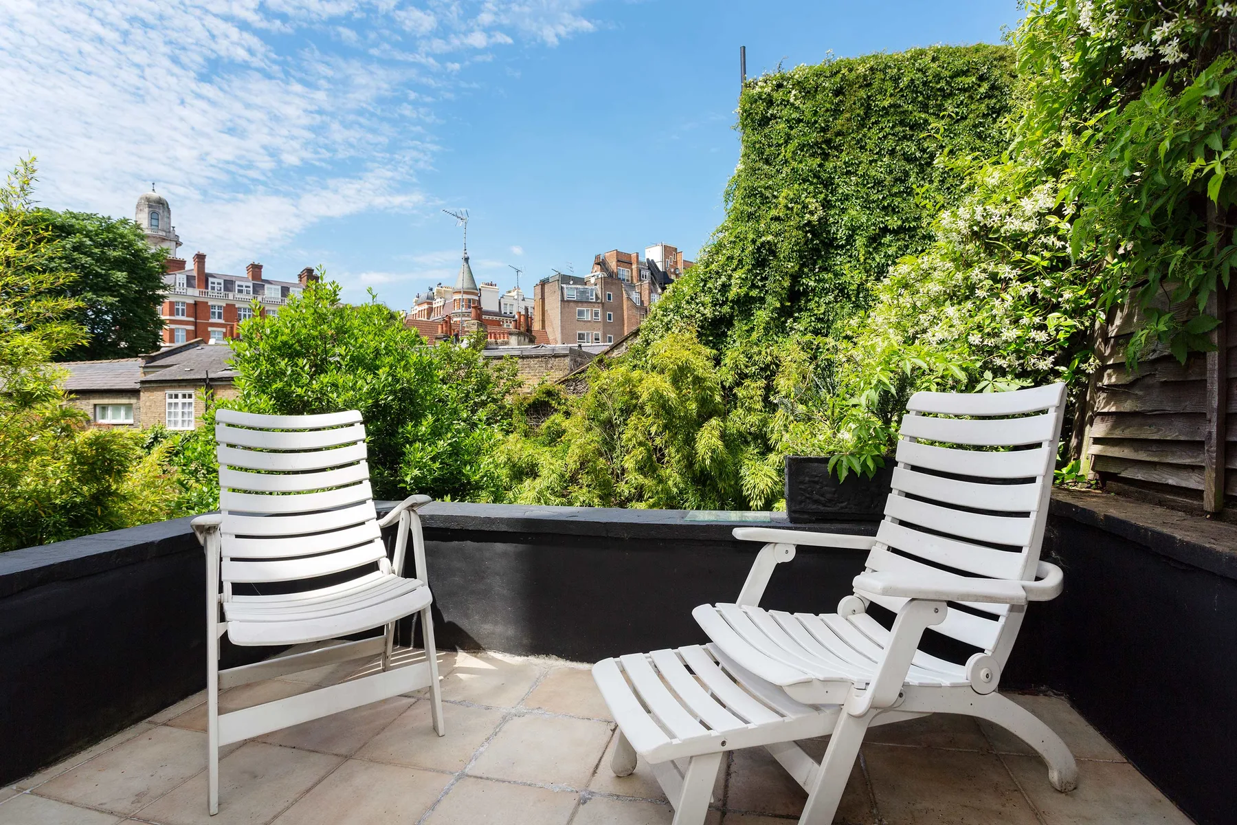 A rooftop terrace with white outdoor seating overlooks leafy greenery and nearby buildings.