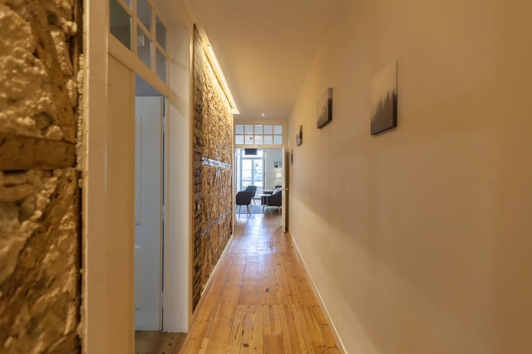 A hallway with wooden flooring leads toward a bright seating area.