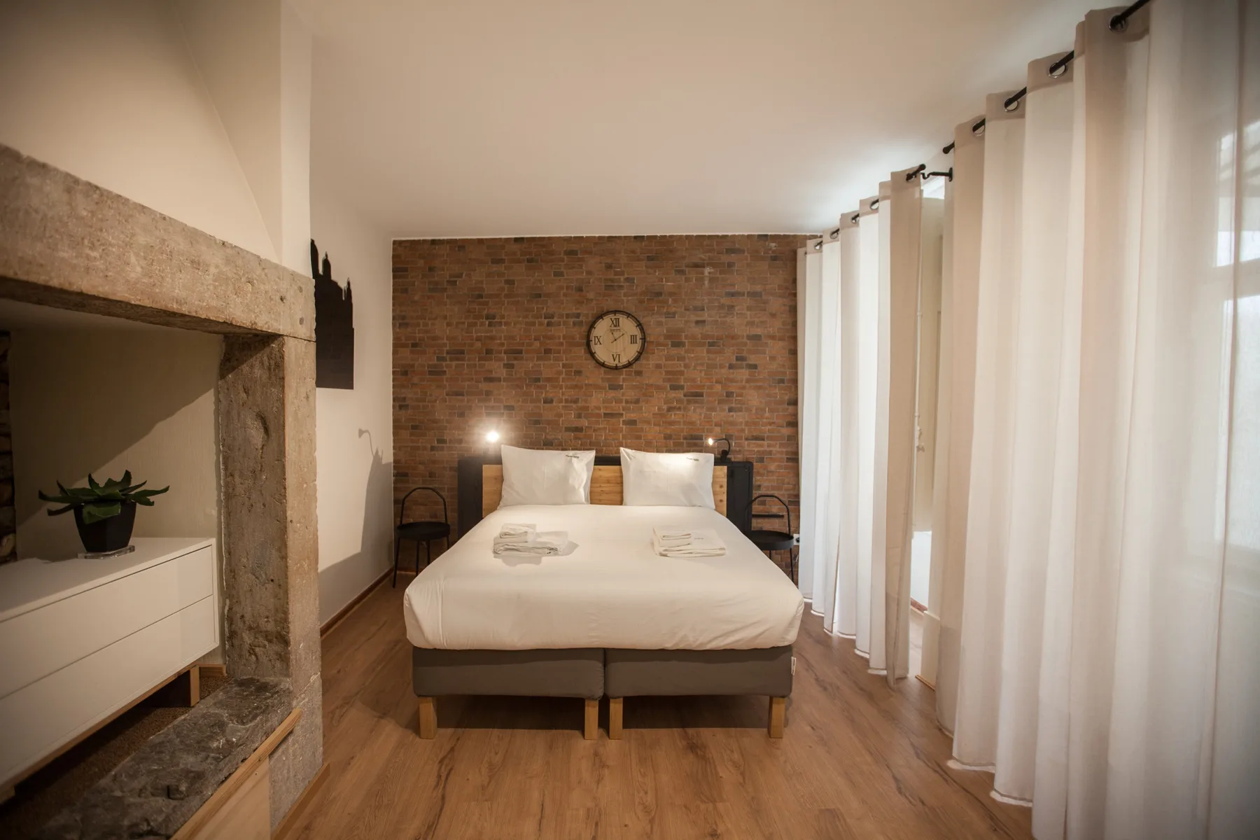 A bedroom with a bed set against a brick wall and curtains along the windows.