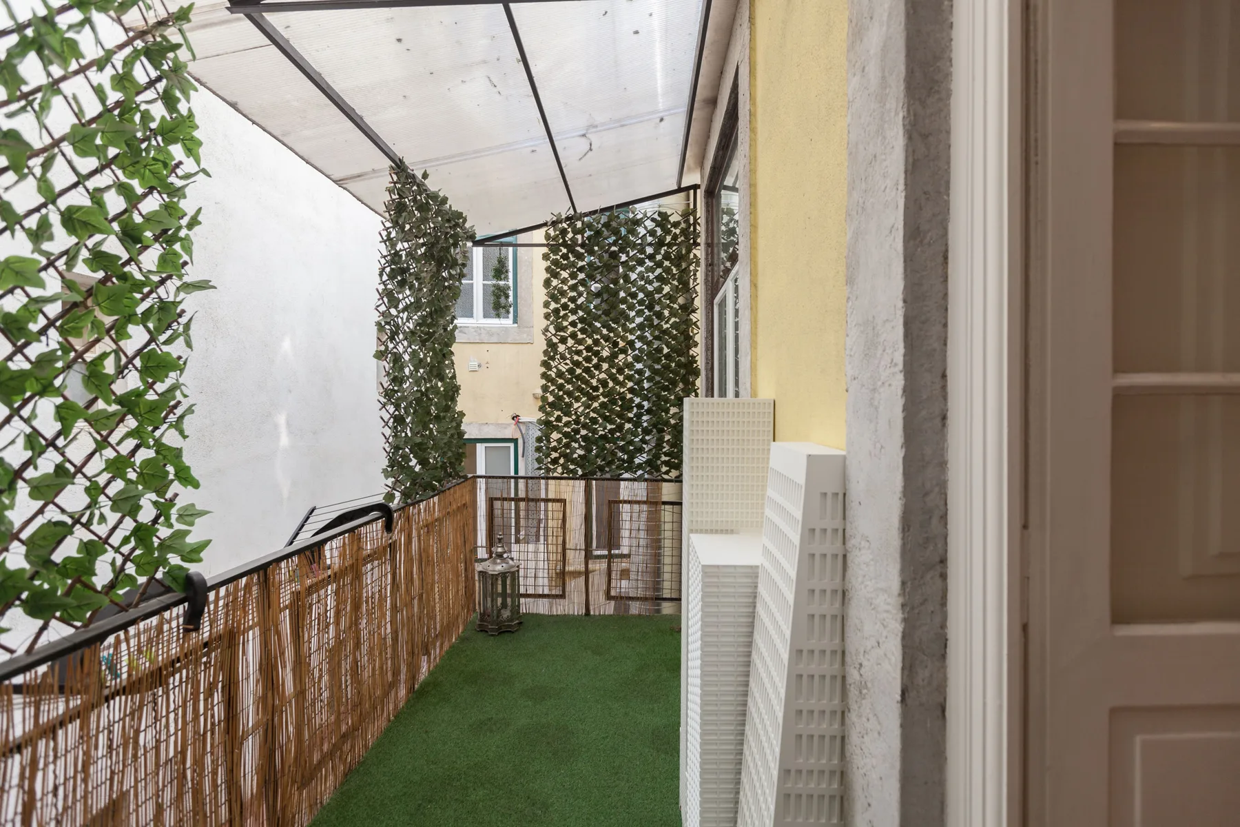 A narrow outdoor terrace with green flooring, climbing plants, and a translucent canopy.