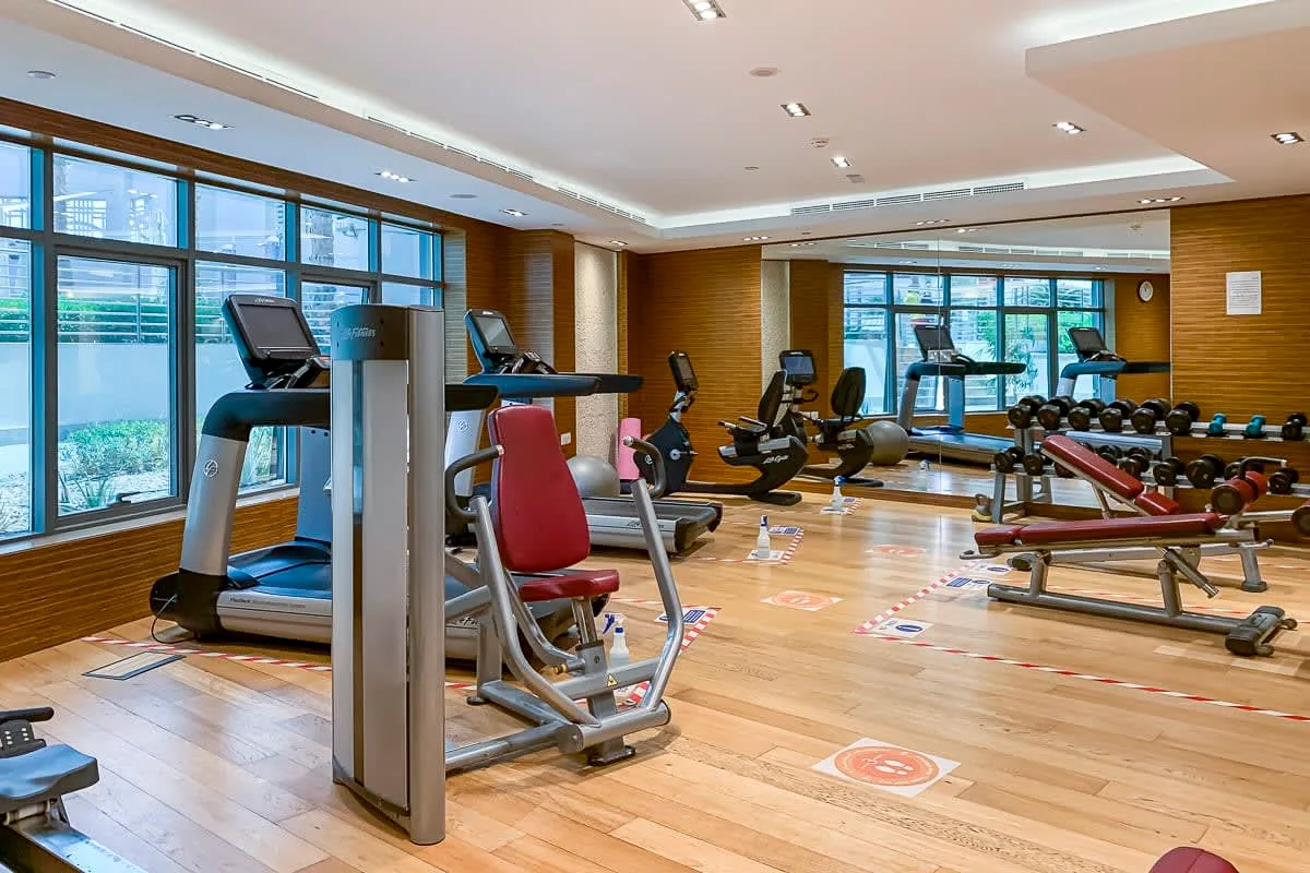 Un gimnasio con máquinas de ejercicio, bancos de pesas y grandes ventanas.