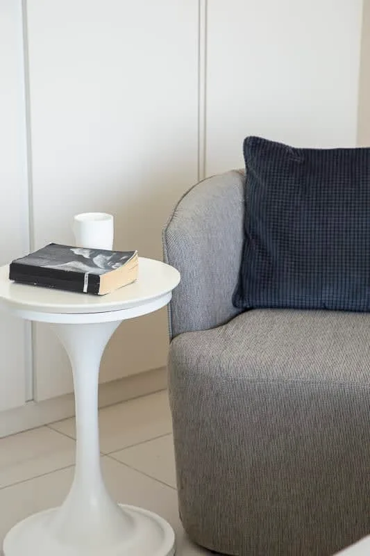 Un asiento tapizado gris con un cojín oscuro está junto a una pequeña mesa blanca.