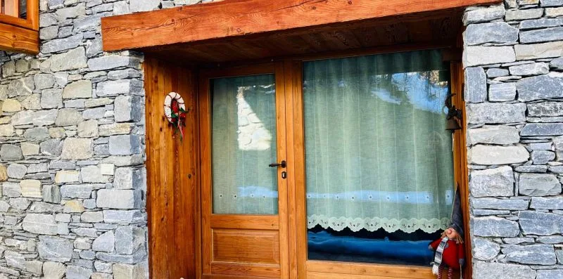 A wooden entrance door and curtained window are set into a stone wall.