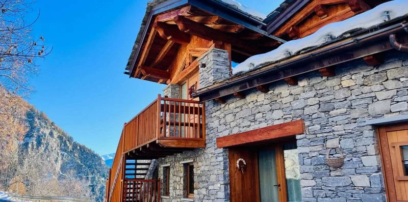 A stone house with wooden balconies stands against a mountain backdrop.
