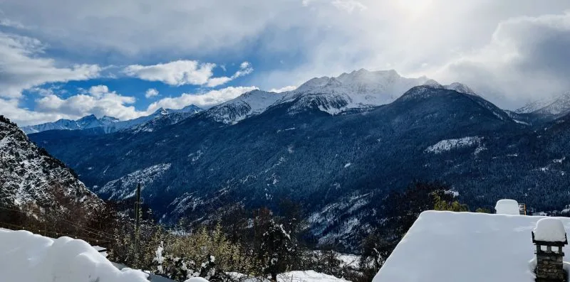 A snowy mountain range rises beyond a snow-covered rooftop beneath a cloudy sky.