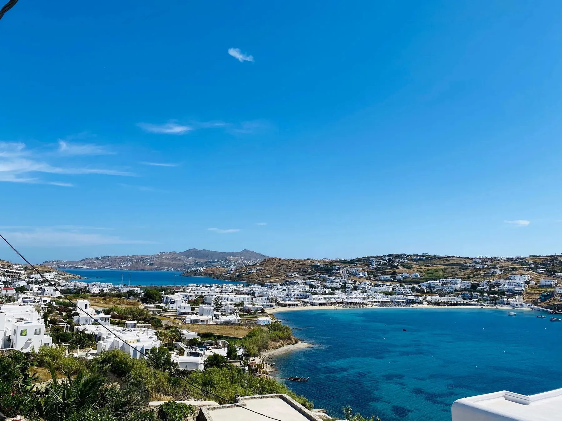 Coastal landscape with blue water, hillside buildings, and distant hills.
