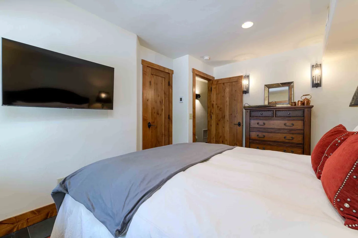 A bedroom with a bed, wall-mounted television, wooden doors, and a dresser.