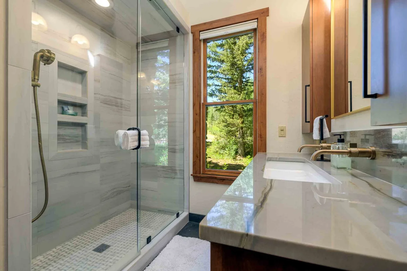 A bright bathroom has a glass shower beside a long counter and window.