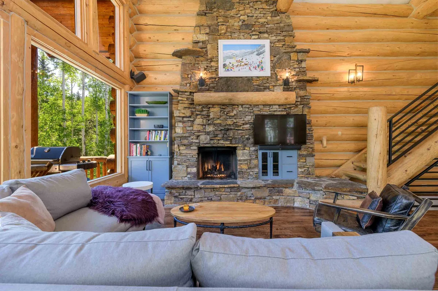 Una sala de estar de cabaña de troncos con chimenea de piedra, televisor y asientos.