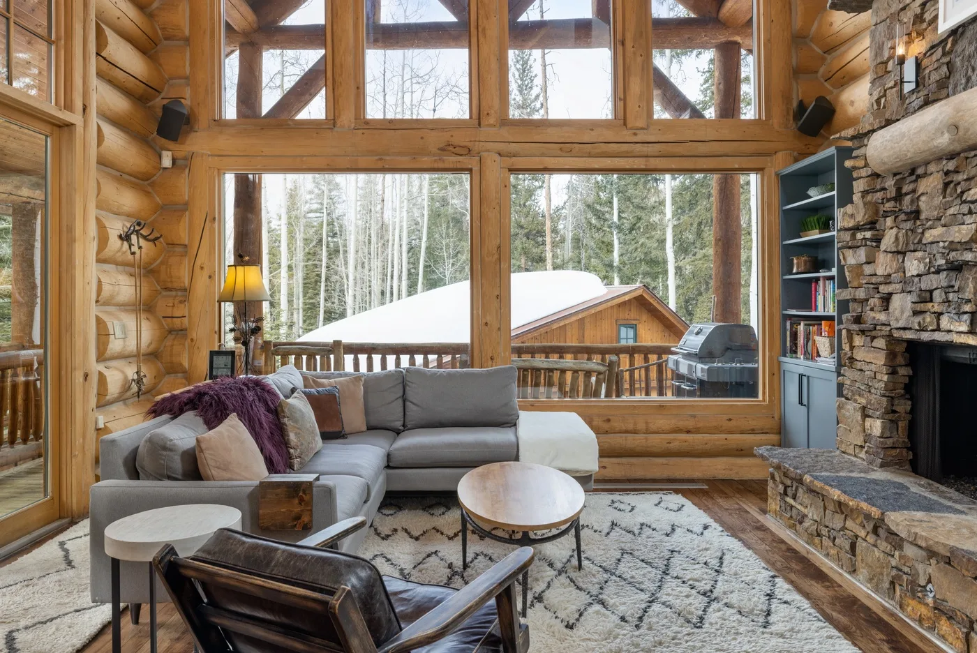 A log cabin living room with seating facing large windows and a stone fireplace.