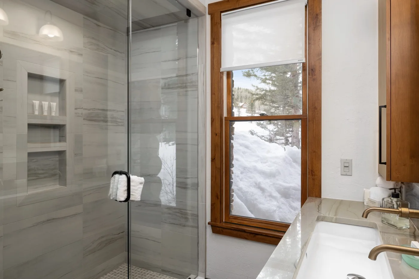 A bathroom with a glass shower, sink, and window overlooking snow.