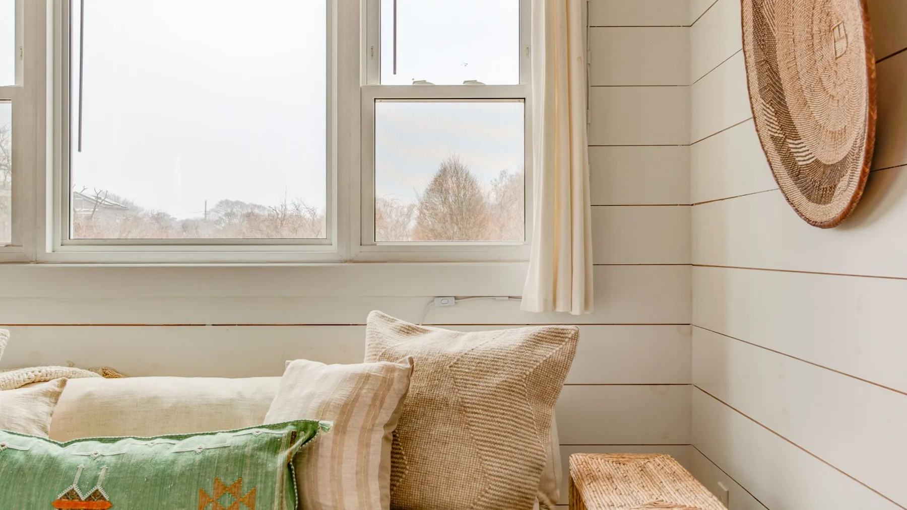 Una zona de estar luminosa junto a grandes ventanas, con cojines y un adorno tejido en la pared.