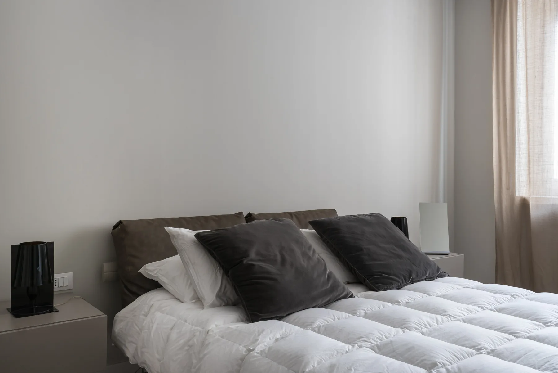 A bedroom with a neatly made bed and dark pillows beside a window.