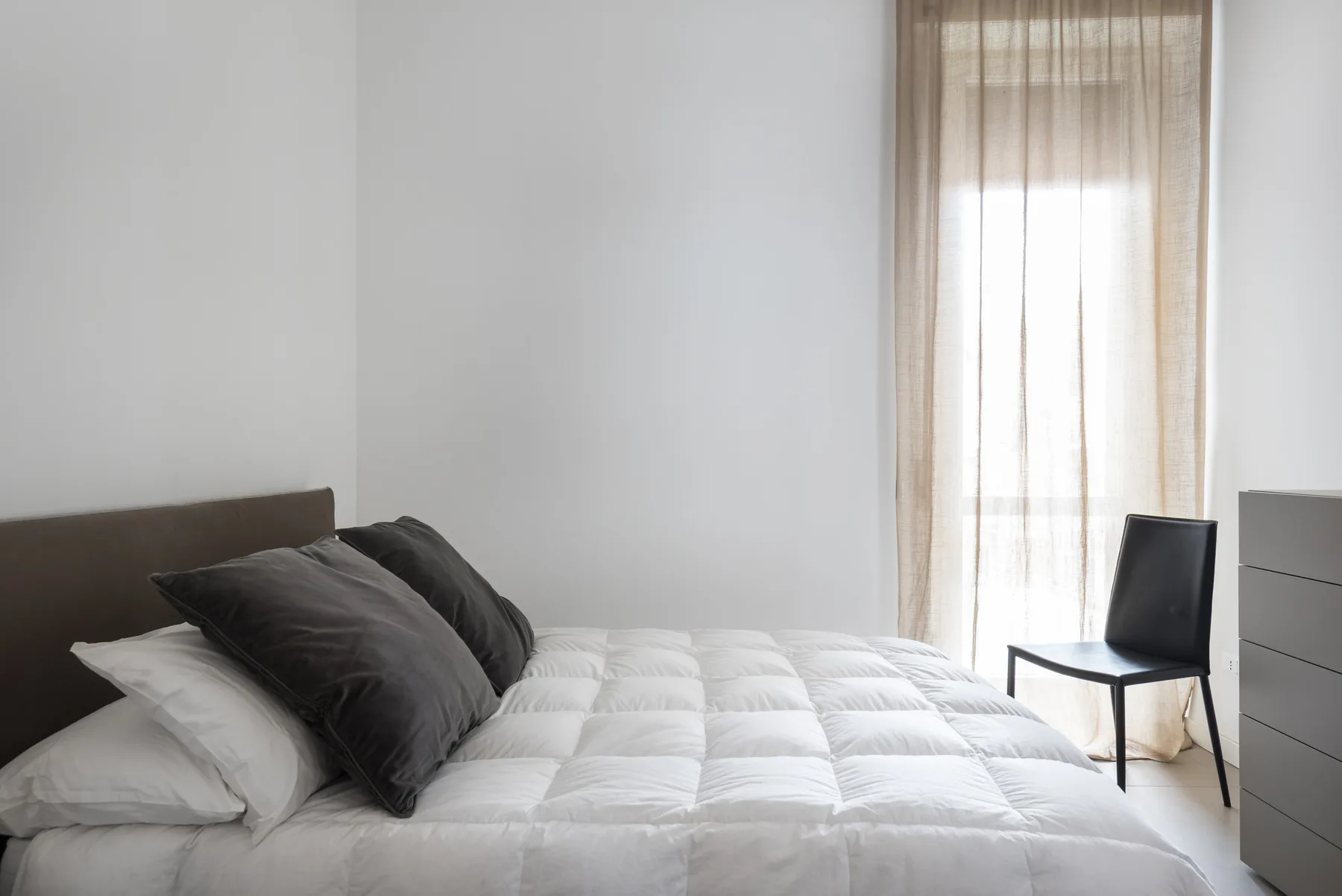 A bright bedroom with a bed dressed in white bedding and dark pillows.
