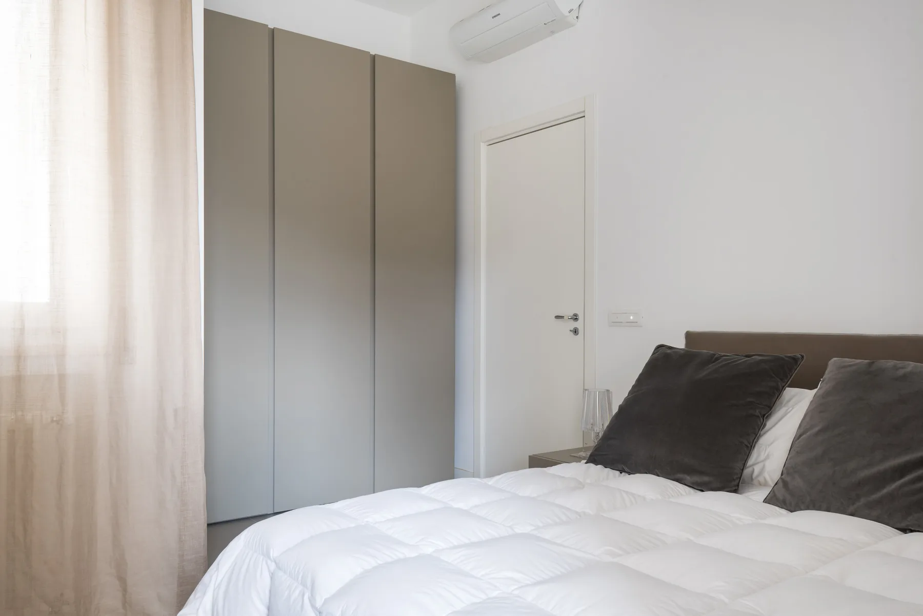 A bright bedroom with a white bed, dark pillows, and a tall wardrobe.