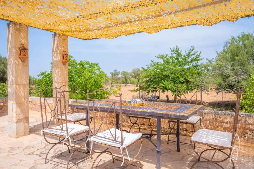Un comedor al aire libre está bajo una cubierta de sombra tejida, rodeado de árboles.