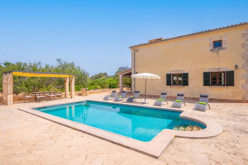 Una piscina exterior está junto a una casa soleada y asientos en el patio.