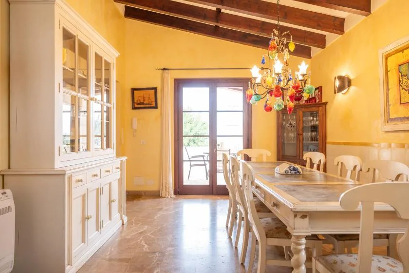 Un comedor con una mesa larga, asientos blancos y una lámpara de araña colorida.