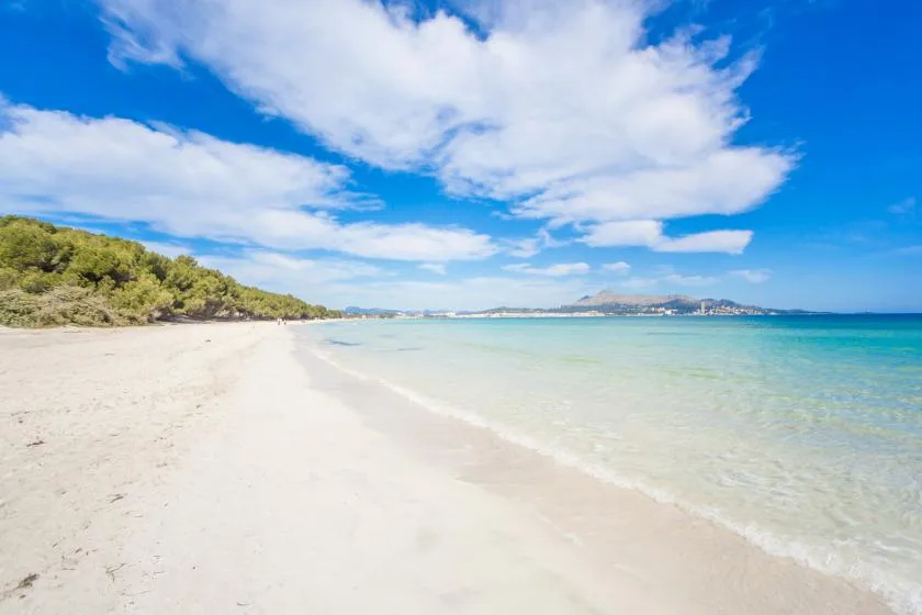 Una playa de arena se curva junto a aguas poco profundas y claras bajo un cielo azul con nubes.