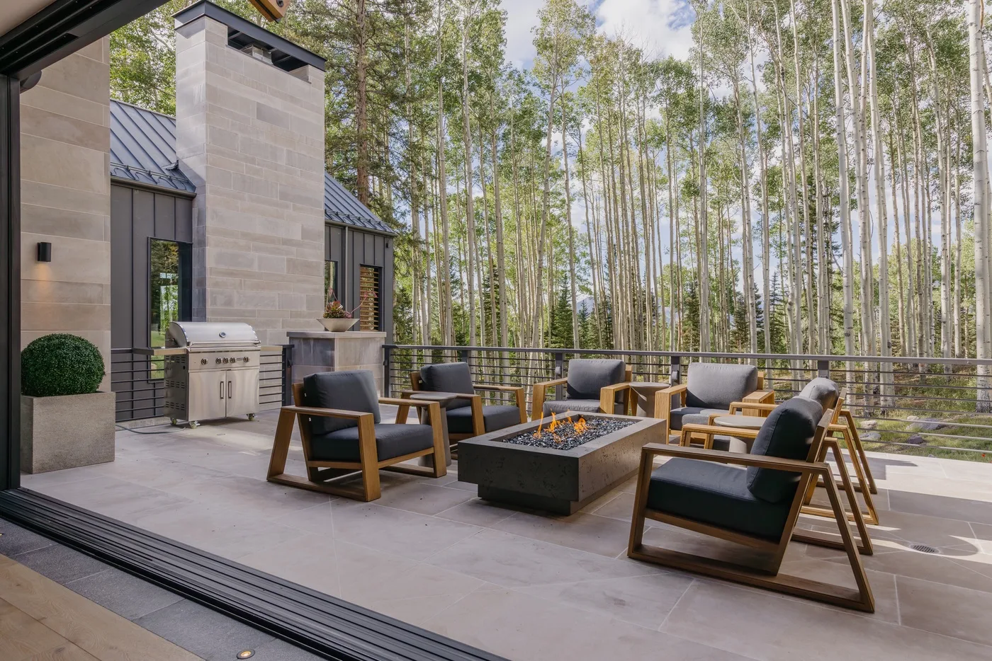 An outdoor patio has lounge seating around a square fire pit beside a wooded backdrop.
