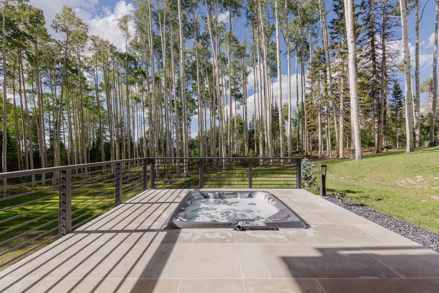 A paved deck with a hot tub overlooks a wooded lawn.