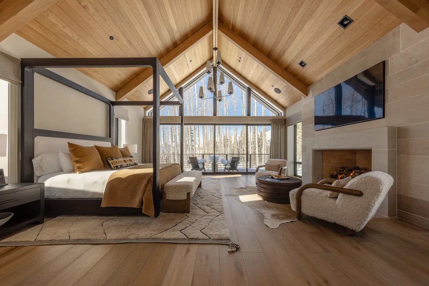 A bedroom and sitting area sit beneath a vaulted wooden ceiling, with windows across the far wall.
