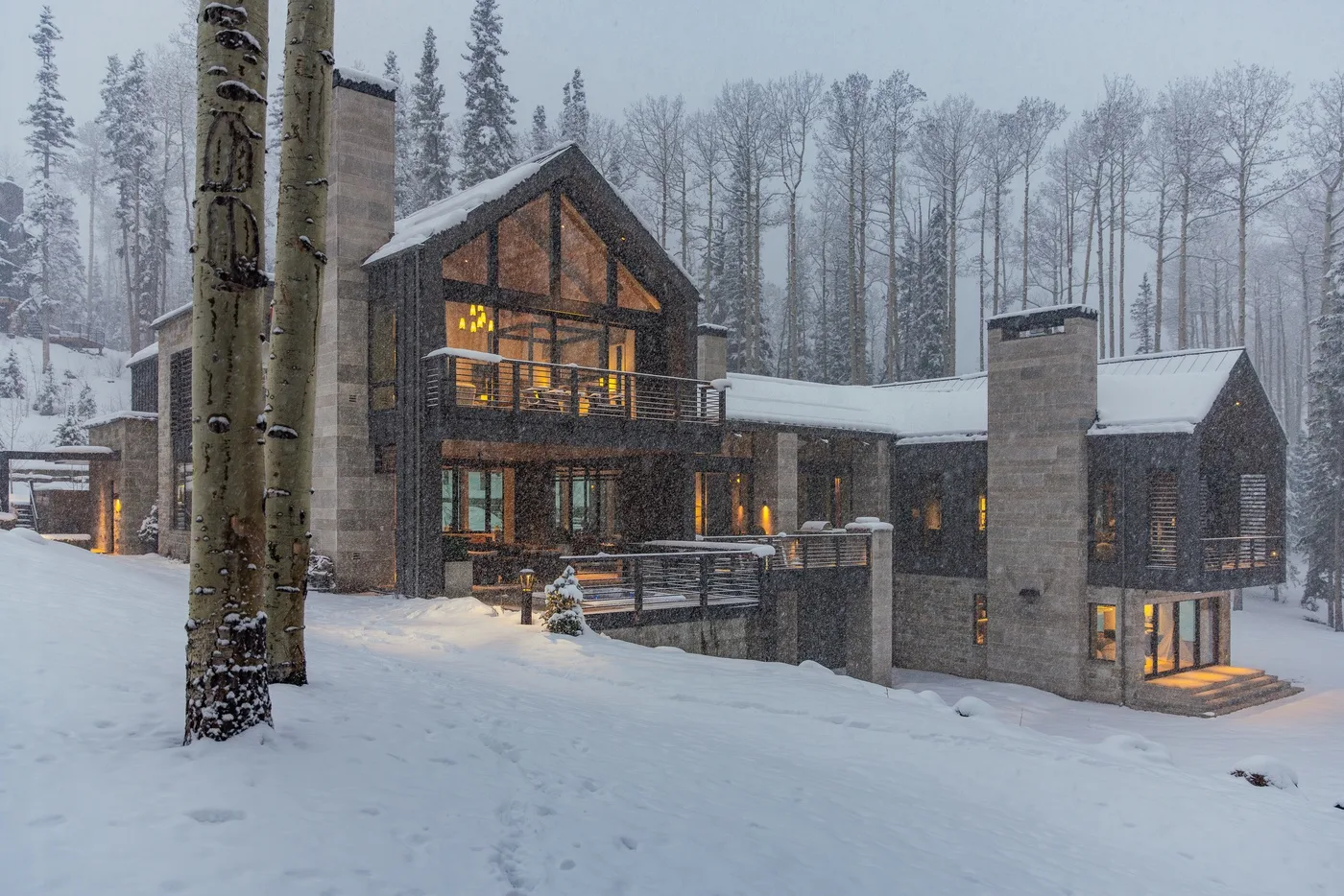 A modern house sits in a snowy forest, with warm lights glowing through its windows.