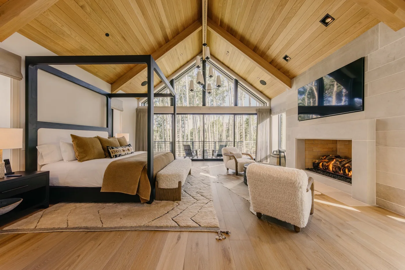 A bedroom and sitting area beneath a vaulted wood ceiling, with a fireplace and large windows.