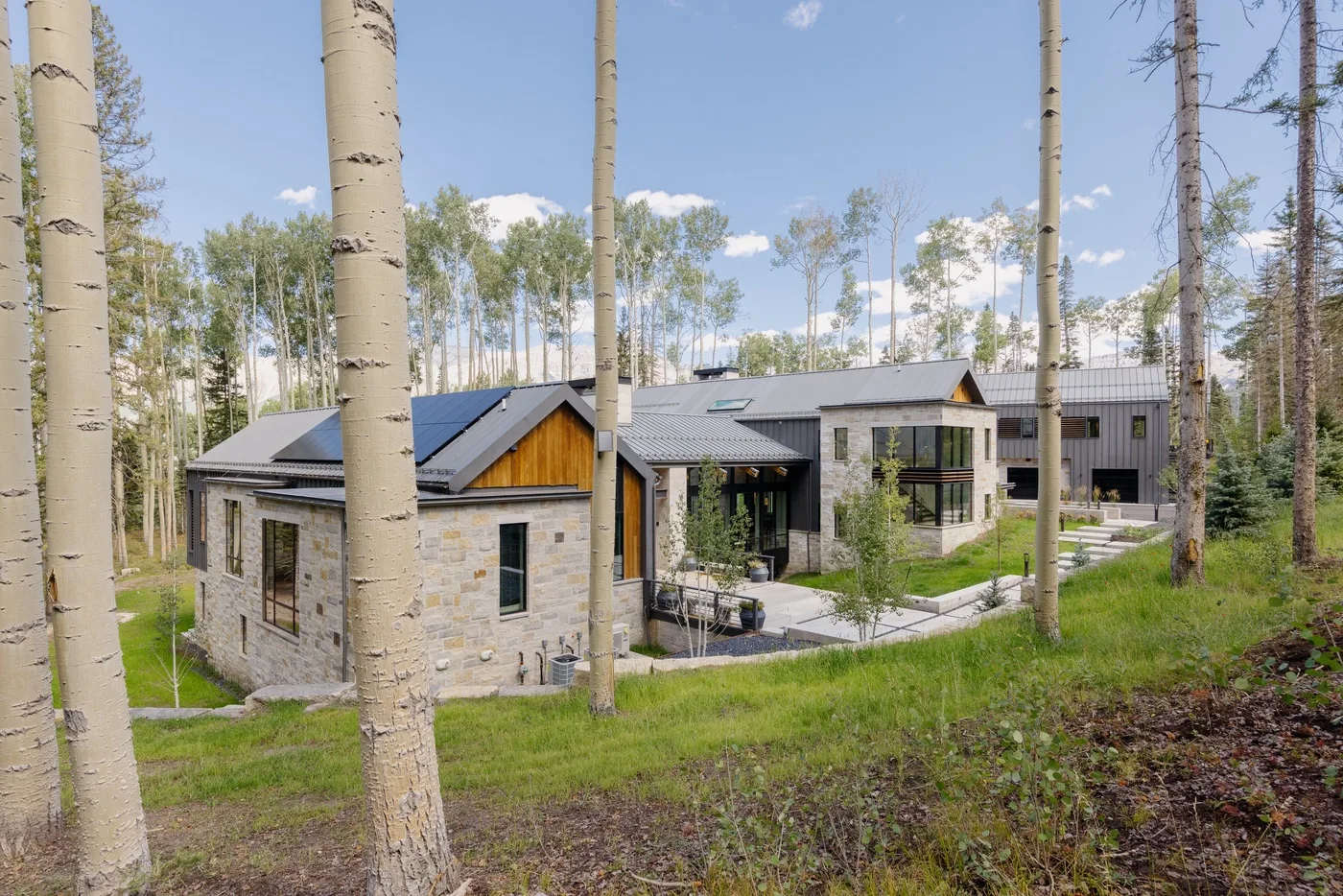 A modern stone house sits among tall trees beside a grassy slope.